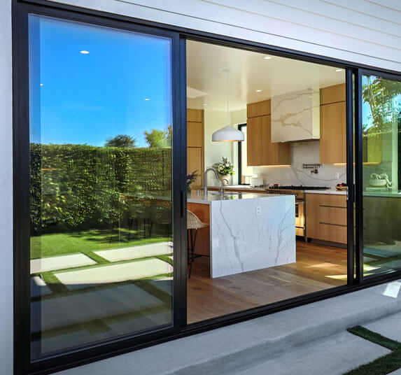Black framed sliding glass door reflecting a hedged backyard while revealing a modern kitchen with a marble island.