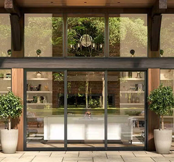 Modern glass sliding door entrance flanked by potted topiaries revealing a fireplace and dining table inside.