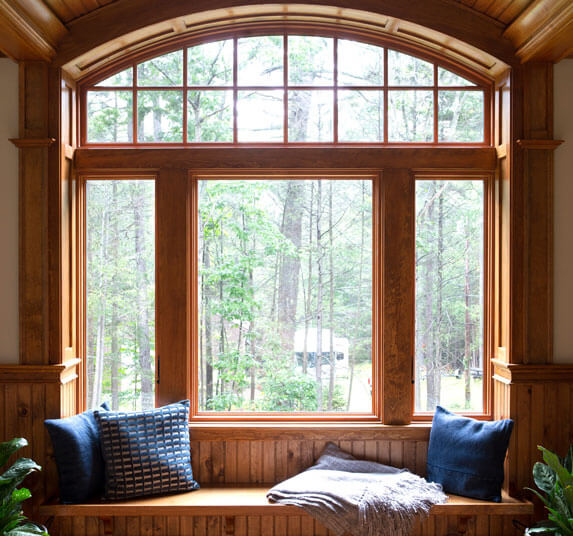 Wood framed arched window seat with blue pillows and a throw blanket overlooking a wooded backyard.