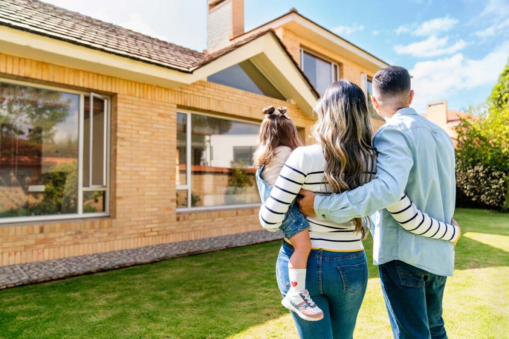 Family embracing while looking toward a brick house with a chimney and gabled roof on a sunny day.